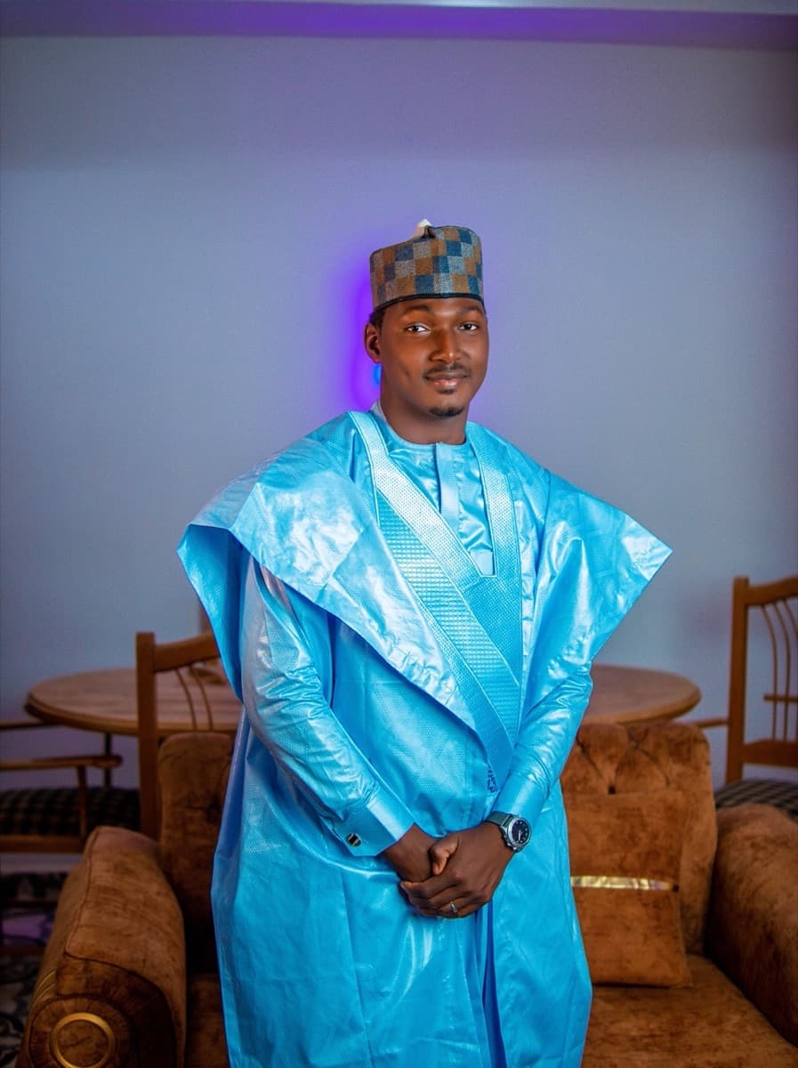 A man wearing a vibrant sky-blue agbada with dramatic wide draped sleeves, an embroidered chest panel, and a patterned cap