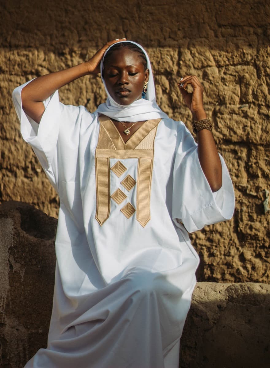 A woman in a structured white oversized robe with a gold geometric embroidered chest panel, against a stone wall
