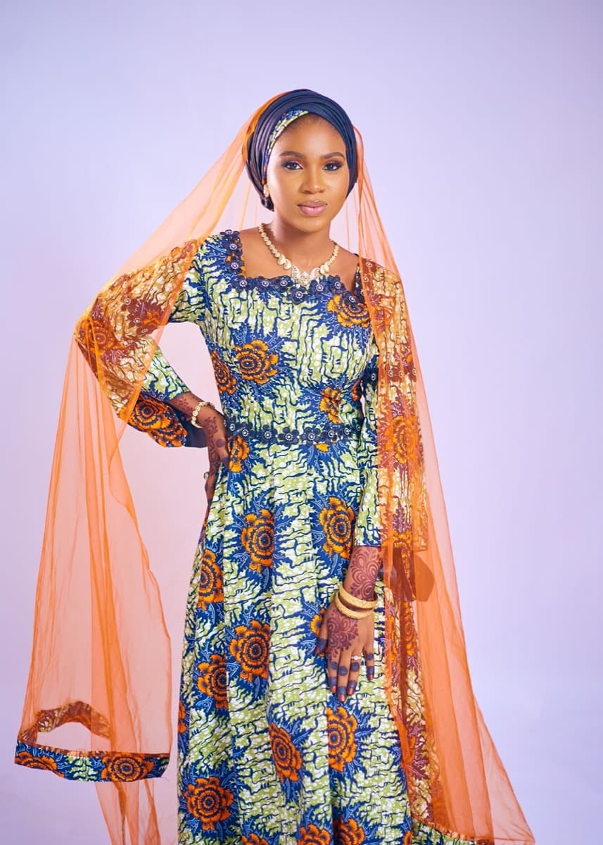 A woman in a flowing blue and orange floral-print ankara gown with a sheer orange veil, hands on hips
