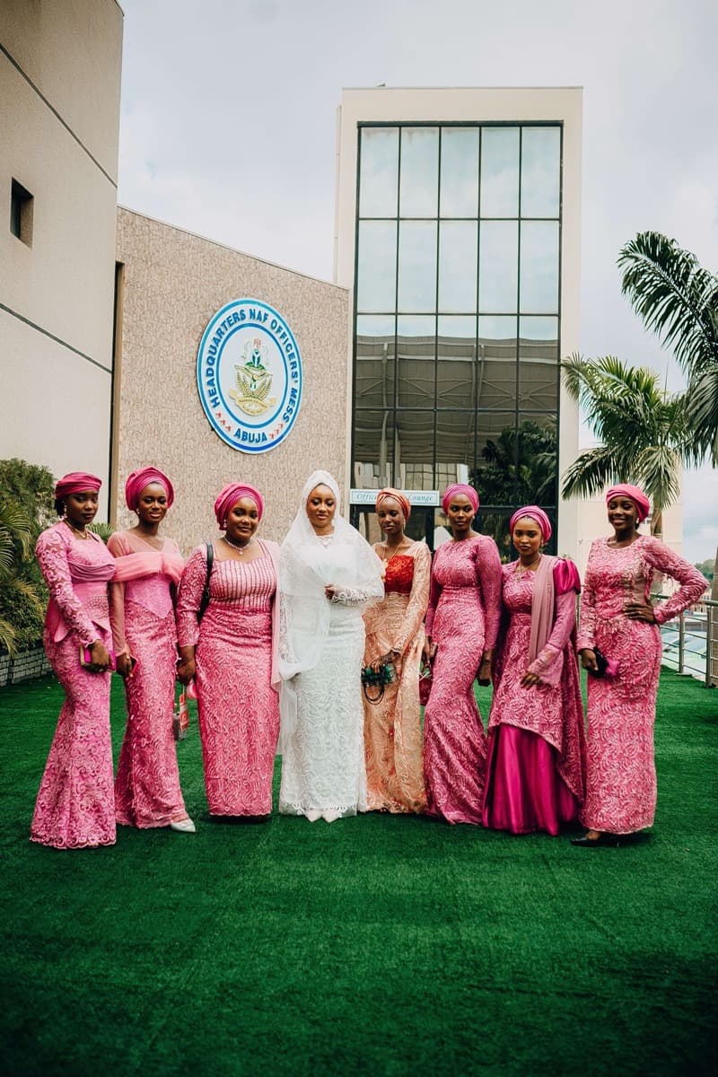 A bride in white standing with bridesmaids in matching pink lace outfits outside a building in Abuja