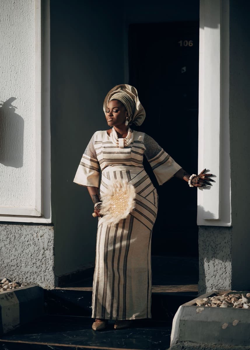 A woman in a fitted cream and brown striped aso oke gown, standing in a doorway holding a woven fan, with a visible mitered seam where the stripe direction changes at the waist