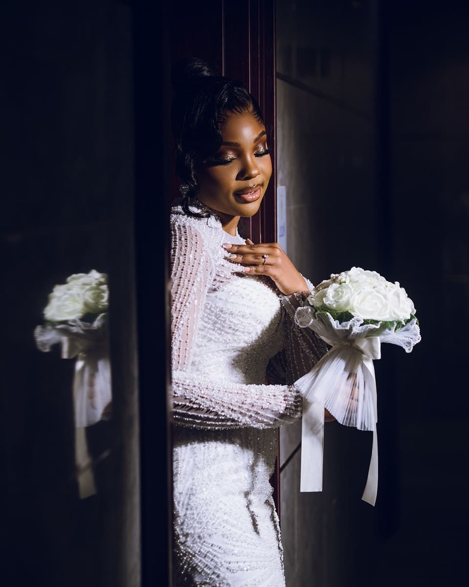 A Nigerian bride in a fitted white beaded long-sleeve gown, holding a white bouquet