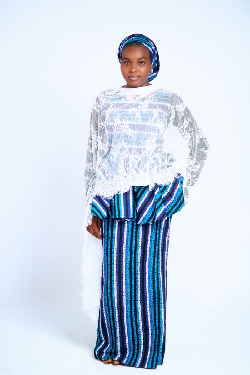 A woman in a white lace buba blouse over a blue and black striped iro wrapper skirt, with a matching blue gele