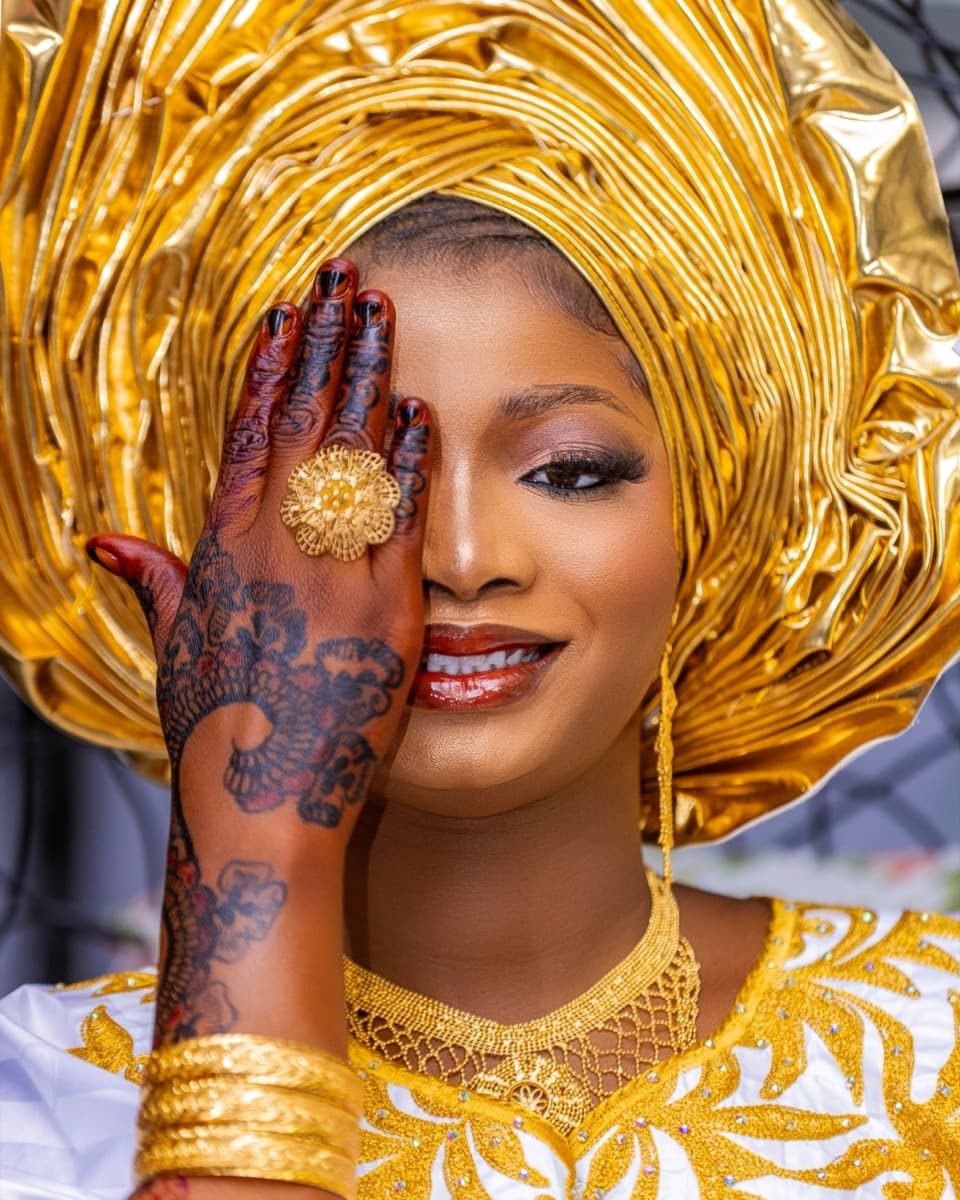 A woman wearing a dramatic gold gele with sweeping pleats, holding up a henna-decorated hand