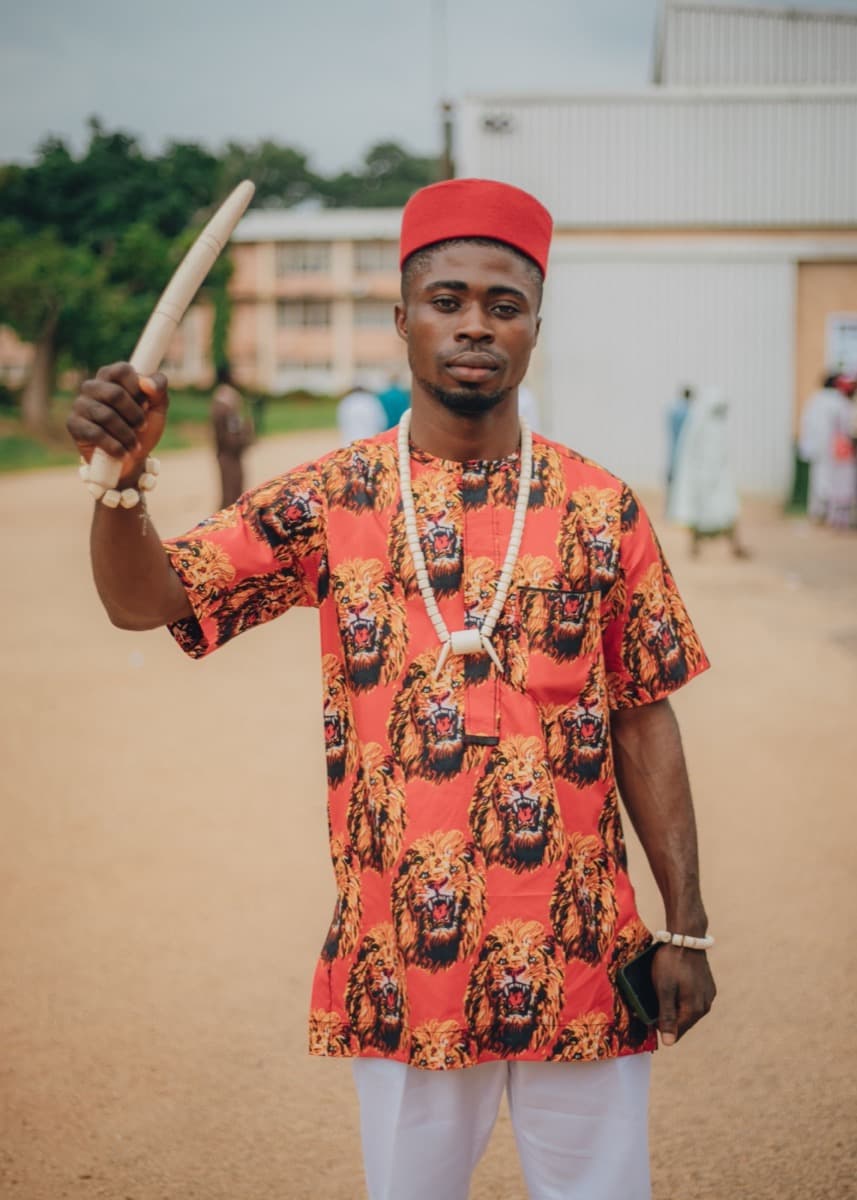 A man outdoors wearing a red isiagu shirt with a repeating gold lion-head print, a matching red cap, and coral beads, holding a carved staff