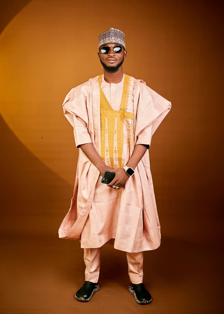 A man wearing a flowing peach babban riga with a gold embroidered chest panel, an embroidered Hausa hula cap, and sunglasses