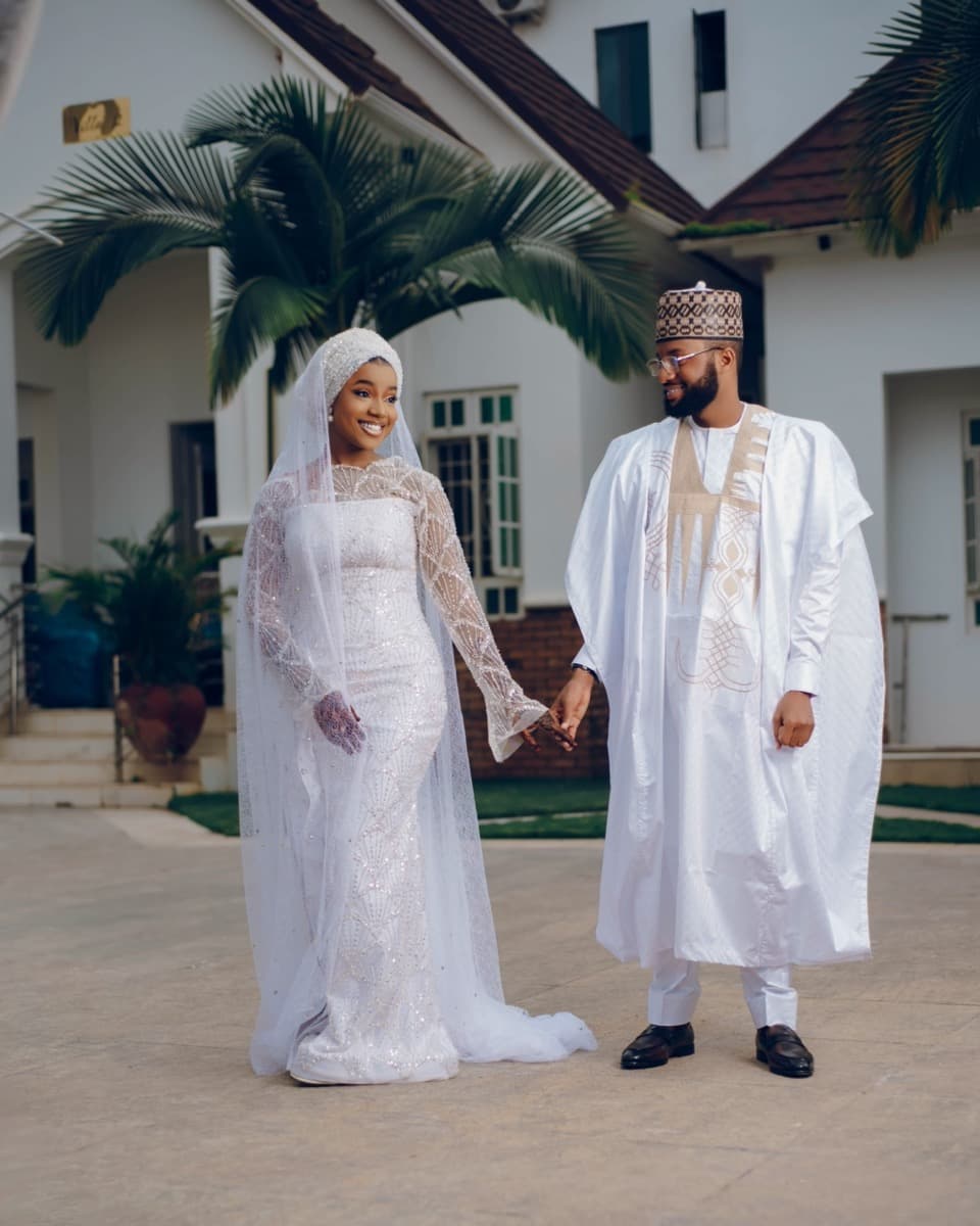 A Nigerian bride in a fitted white lace gown and veil standing hand-in-hand with a groom in white agbada