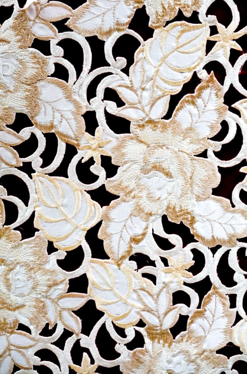Close-up of cream and gold guipure lace with raised, corded floral and leaf motifs against a black background