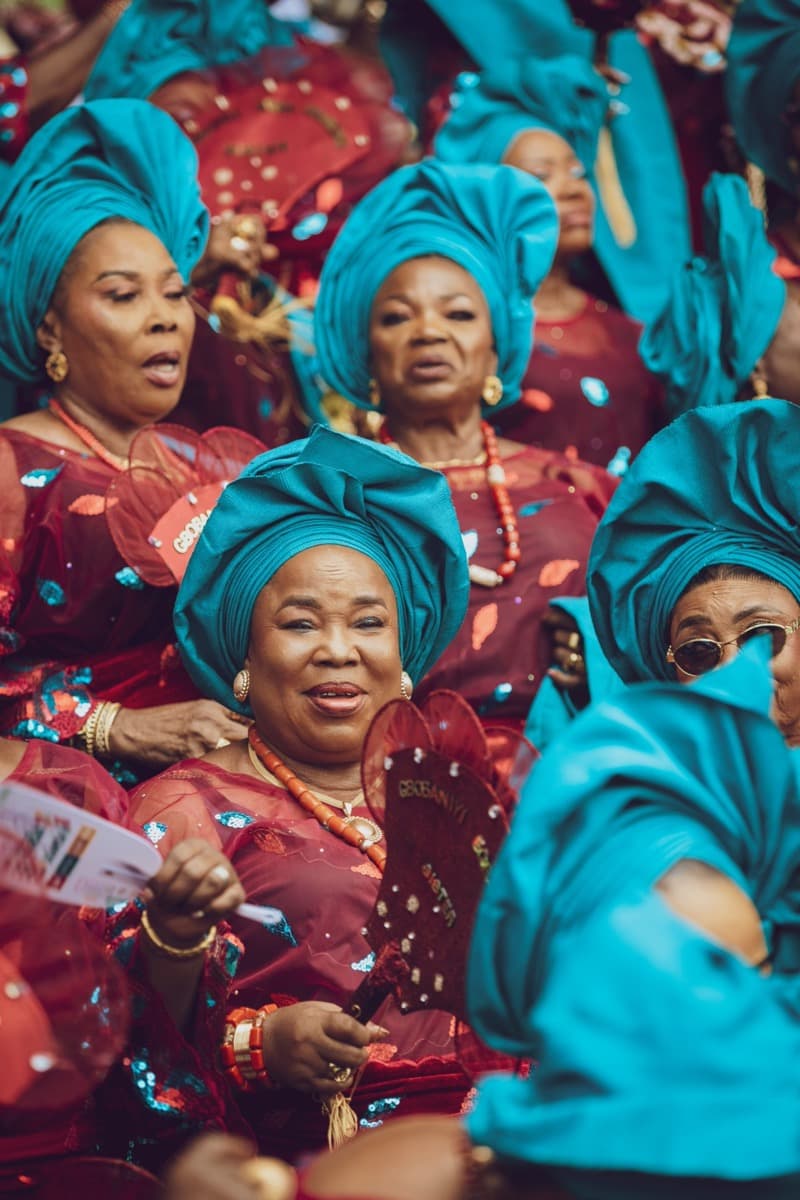A crowd of women in matching red aso-ebi and teal geles at a Nigerian celebration, holding event fans with confetti in the air