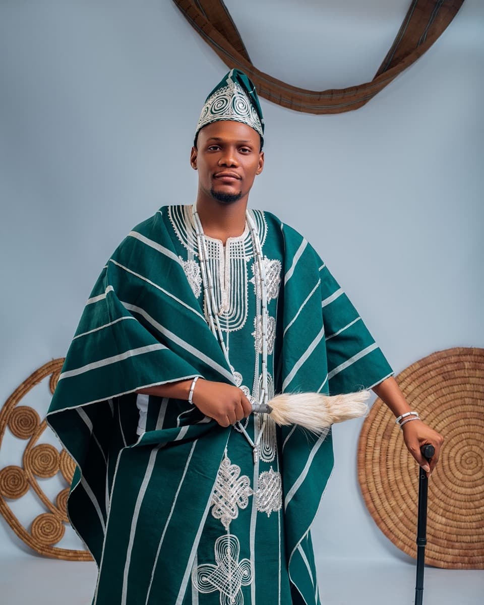 A man in a deep green agbada with extensive silver embroidery, an embroidered fila cap, holding a horsetail fly-whisk and cane