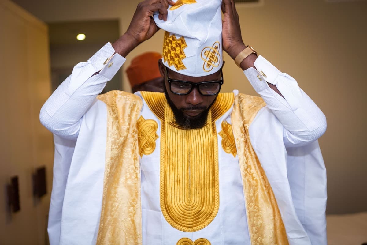 Close-up of a man adjusting his cap, wearing a white agbada with an elaborate gold embroidered chest panel