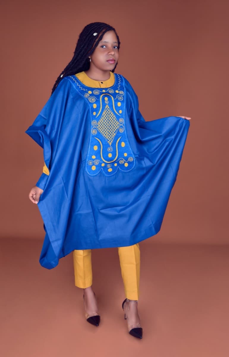 A woman in a vivid blue oversized robe with gold medallion embroidery, worn over fitted mustard-yellow trousers