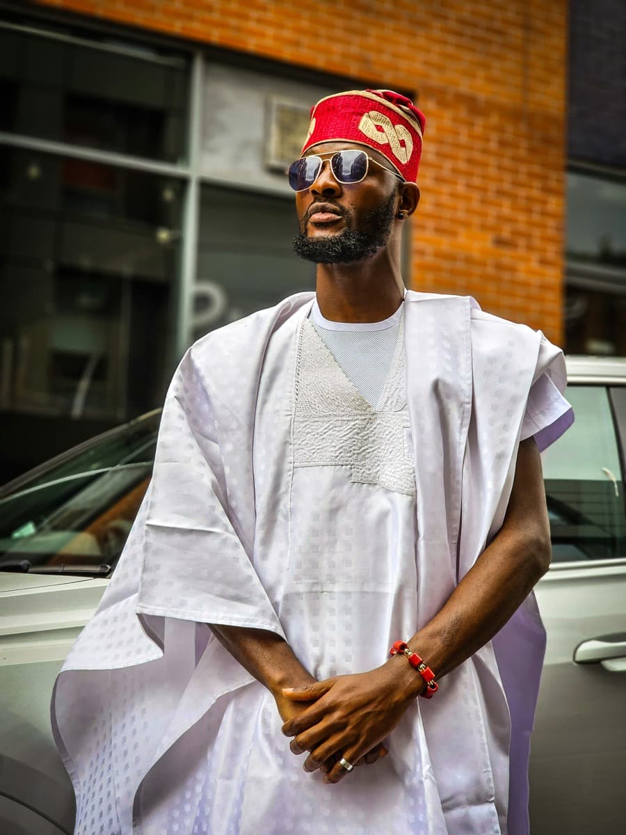 A man in a white and lilac agbada with the sleeves folded back for a slimmer, more tailored look, wearing a red cap and sunglasses