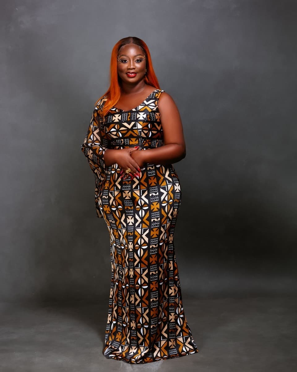 A woman in a fitted brown, orange, and cream geometric-print ankara gown that flares at the hem