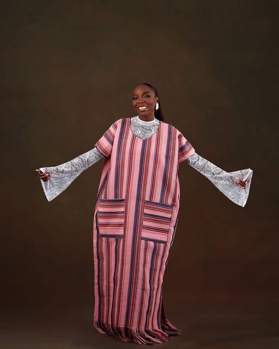 A woman in a wide, flowing pink and black striped aso oke boubou with lace bell sleeves and a lace collar, arms spread to show the loose, robe-like cut