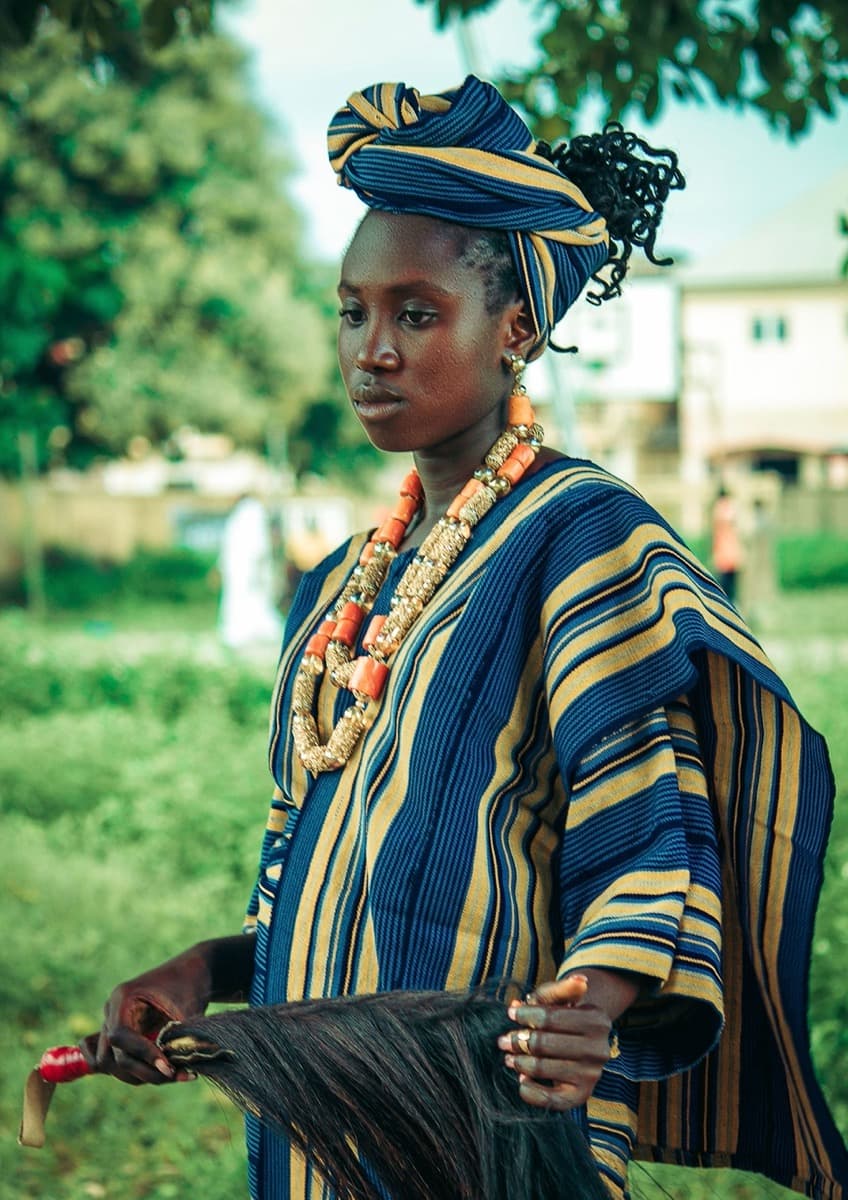 A woman wearing a structured blue and gold striped aso oke wrap top with a matching gele headwrap and layered coral bead necklaces