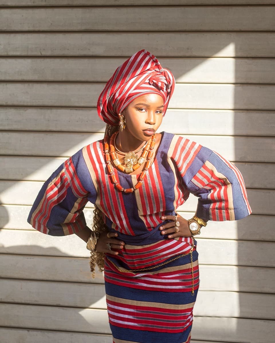 A woman wearing a red, blue, and gold striped aso oke buba blouse cinched with a gold chain belt over a matching iro wrapper skirt, with a matching gele