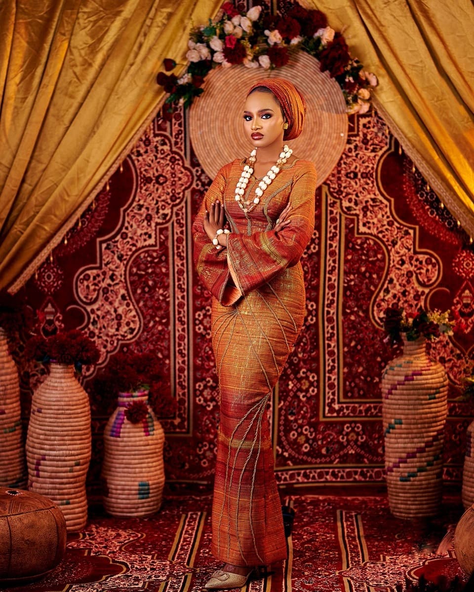 A woman in a fitted, floor-length rust and orange aso oke gown with bell sleeves and a matching gele, standing against a richly patterned backdrop