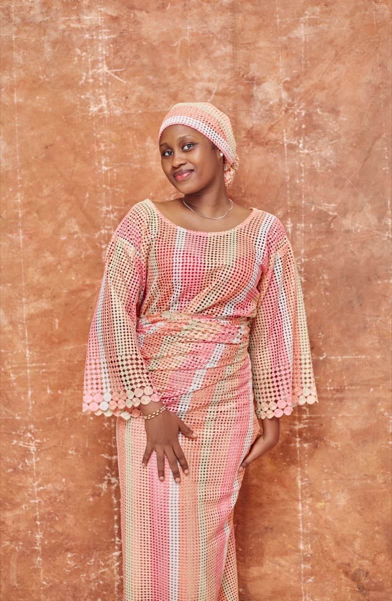 A woman in a loose pink, coral, and gold crochet-lace buba with bell sleeves, gathered at the waist into a matching striped crochet-lace iro wrapper