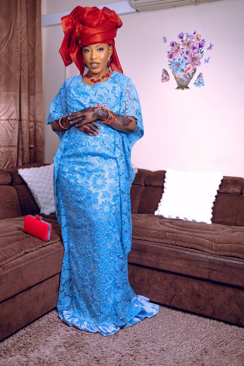A woman in a cornflower-blue floral lace bubu gown with loose kimono-style sleeves and a dramatic red gele