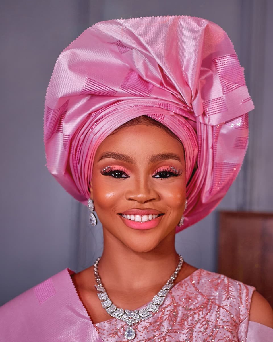 A woman wearing a tall, glossy pink satin gele tied with a bow-like fan structure at the front, with defined sharp pleats