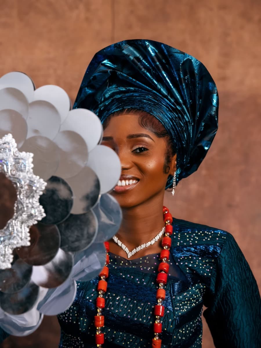 A woman wearing a dark teal velvet gele with clear structured pleating, holding a decorative hand fan