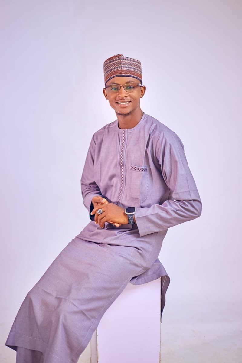 A man seated, wearing a fitted lavender senator-style kaftan with a structured mandarin collar and an embroidered hula cap