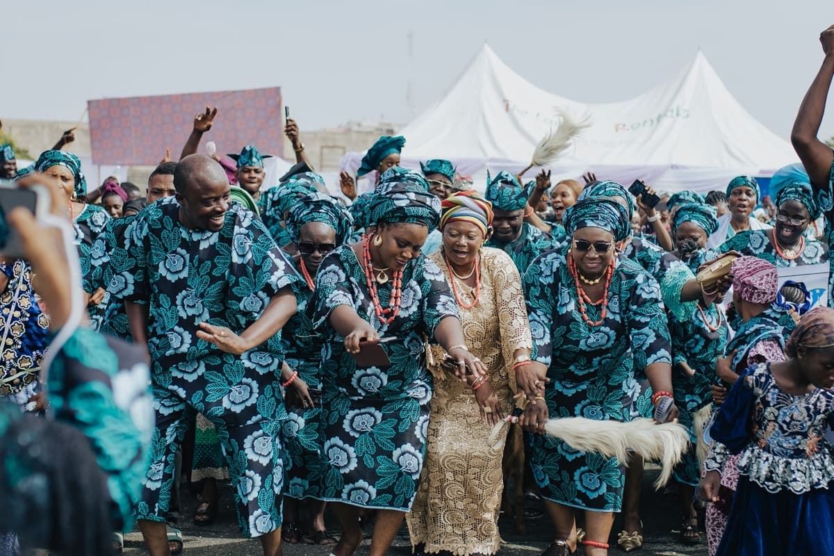 A large crowd dancing outdoors in matching teal ankara prints and coral beads at a Nigerian celebration